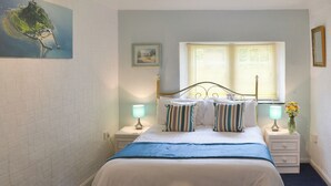A bedroom with a bed, two bedside tables, a window with blinds, a wall-mounted picture, and a nightstand with a lamp. - Rosecraddoc Manor - Natalie Jane (Near Liskeard)