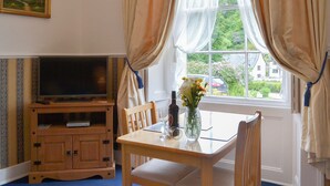 A room with a television on a wooden stand, a table with chairs, a vase with flowers, and a window with curtains. - Rosecraddoc Manor - Natalie Jane (Near Liskeard)