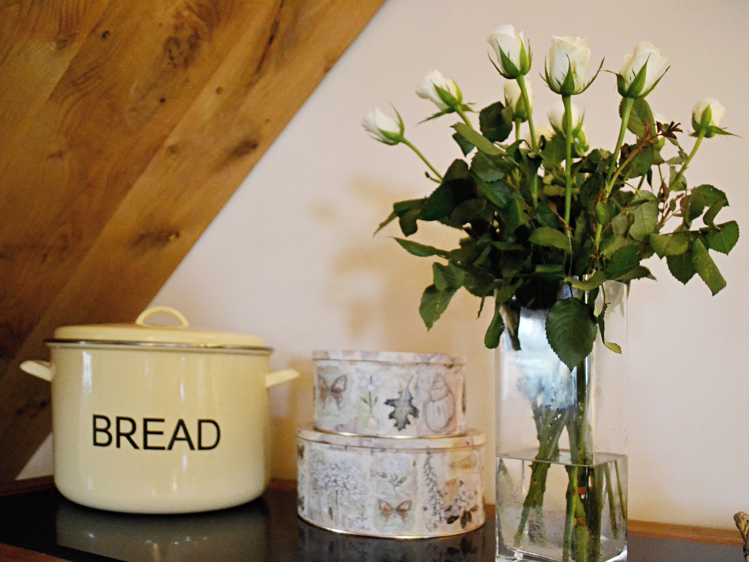 Eine Küchenarbeitsplatte mit einem weißen Topf mit der Aufschrift 'BREAD', einem Behälter mit floralem Muster und einer Vase mit weißen Blumen.