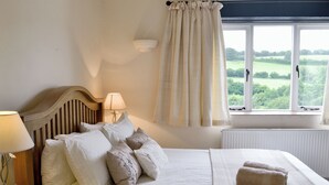 Quarto com cama, almofadas, mesa de cabeceira com abajur, janela com cortinas e vista para o verde lá fora.