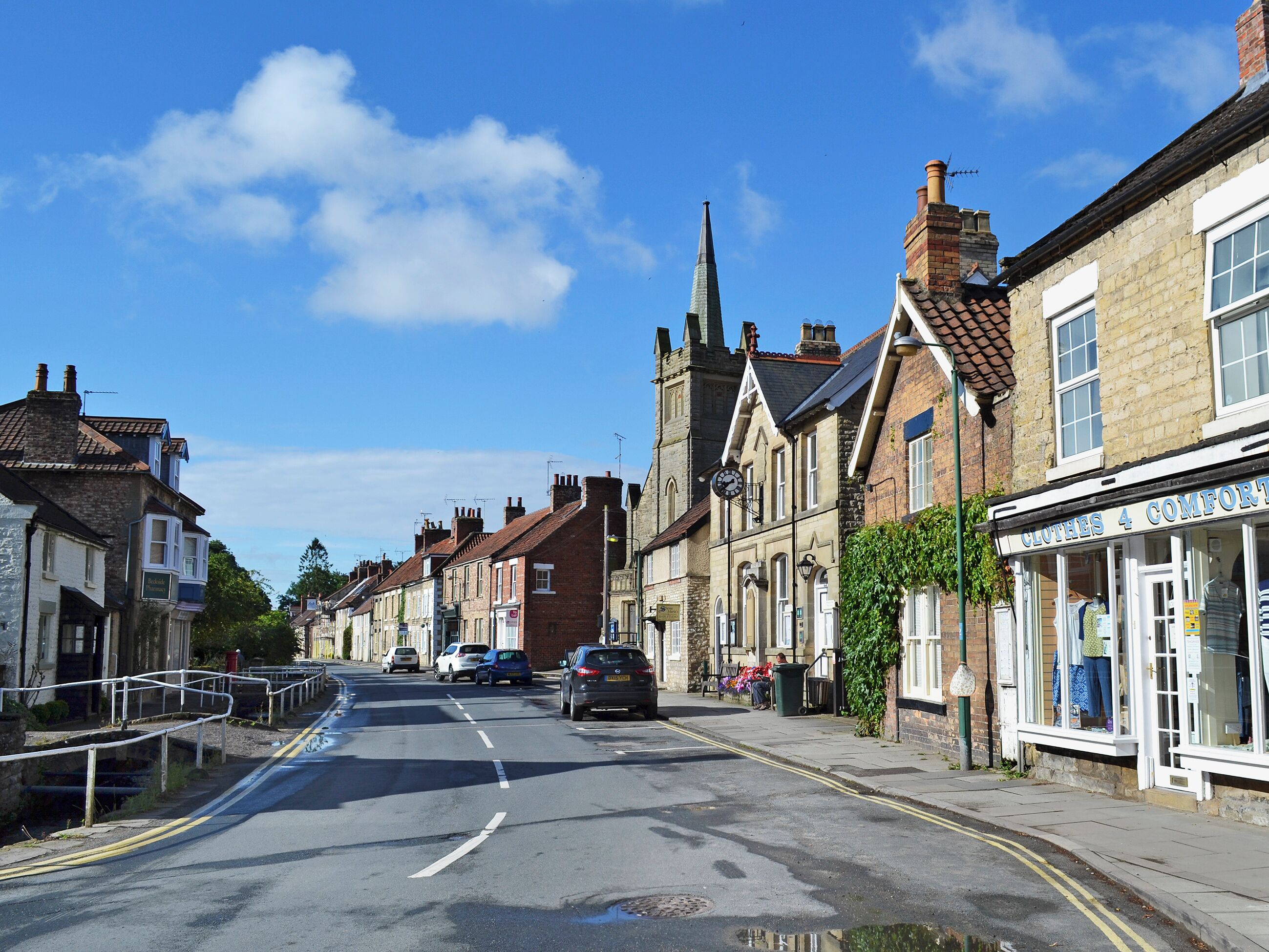 A street with historic buildings, a shop named 'Clothes 4 Comfort', and a church spire in the background.