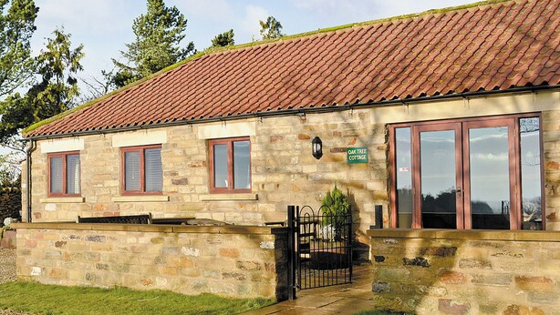 Una cabaña de piedra con techo de tejas, un cartel que dice "Oak Tree Cottage" y una entrada con portón.