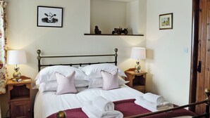 Una cama bien hecha con ropa de cama blanca y almohadas rosadas, una mesita de noche de madera con una lámpara, un espejo sobre la cama y una estantería con una pequeña estatua y un tracto pequeño.
