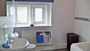 Kylpyhuoneessa on valkoinen pesuallas, wc ja kylpyamme. Pesualtaan yläpuolella on ikkuna ja seinällä pyyheteline, jossa on pyyhkeitä.