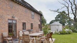 En murstensbygning med en patiohave med et træbord og stole, omgivet af en velplejet græsplæne og have.