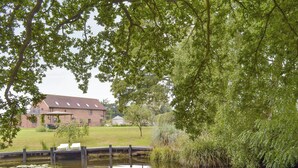 Fredfyldt udsigt over en flod med en murstensbygning, en træbro og frodig vegetation.
