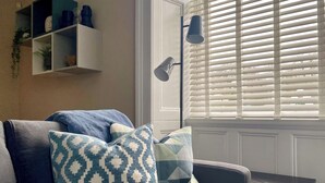 A living room with a grey sofa, patterned cushions, a white wall, a shelf with decorative items, and a window with blinds. - 2 bedroom accommodation in Portinscale, near Keswick (Portinscale, near Keswick)