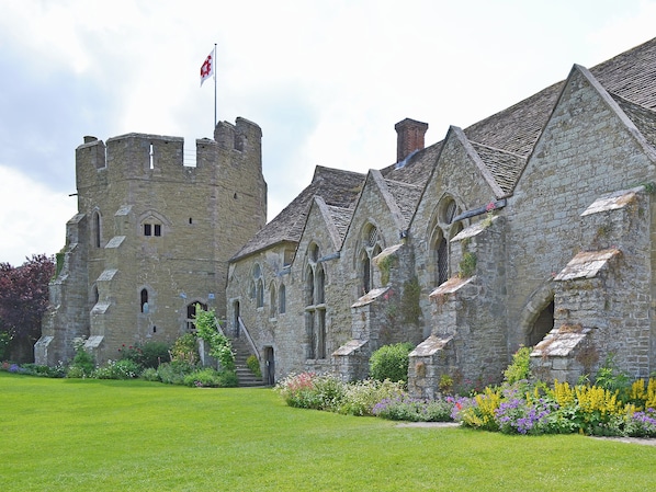 A historic stone building with a flag, featuring a large lawn and colorful flower beds.