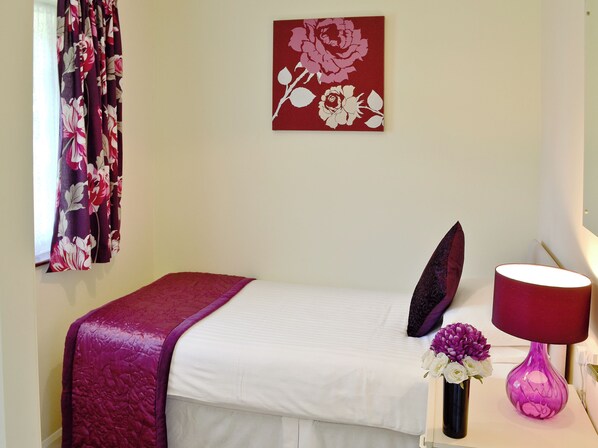 A neatly made bed with a white sheet and a purple blanket, a red and white floral painting on the wall, a vase with flowers, and a lamp on a bedside table.