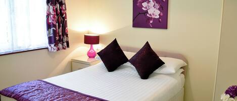 A neatly made bed with a purple bedspread and two dark pillows, a pink lamp on a bedside table, and a purple orchid painting on the wall.