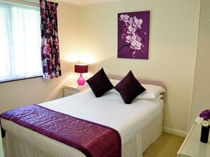 A neatly made bed with a purple bedspread and two dark pillows, a pink lamp on a bedside table, and a purple orchid painting on the wall.