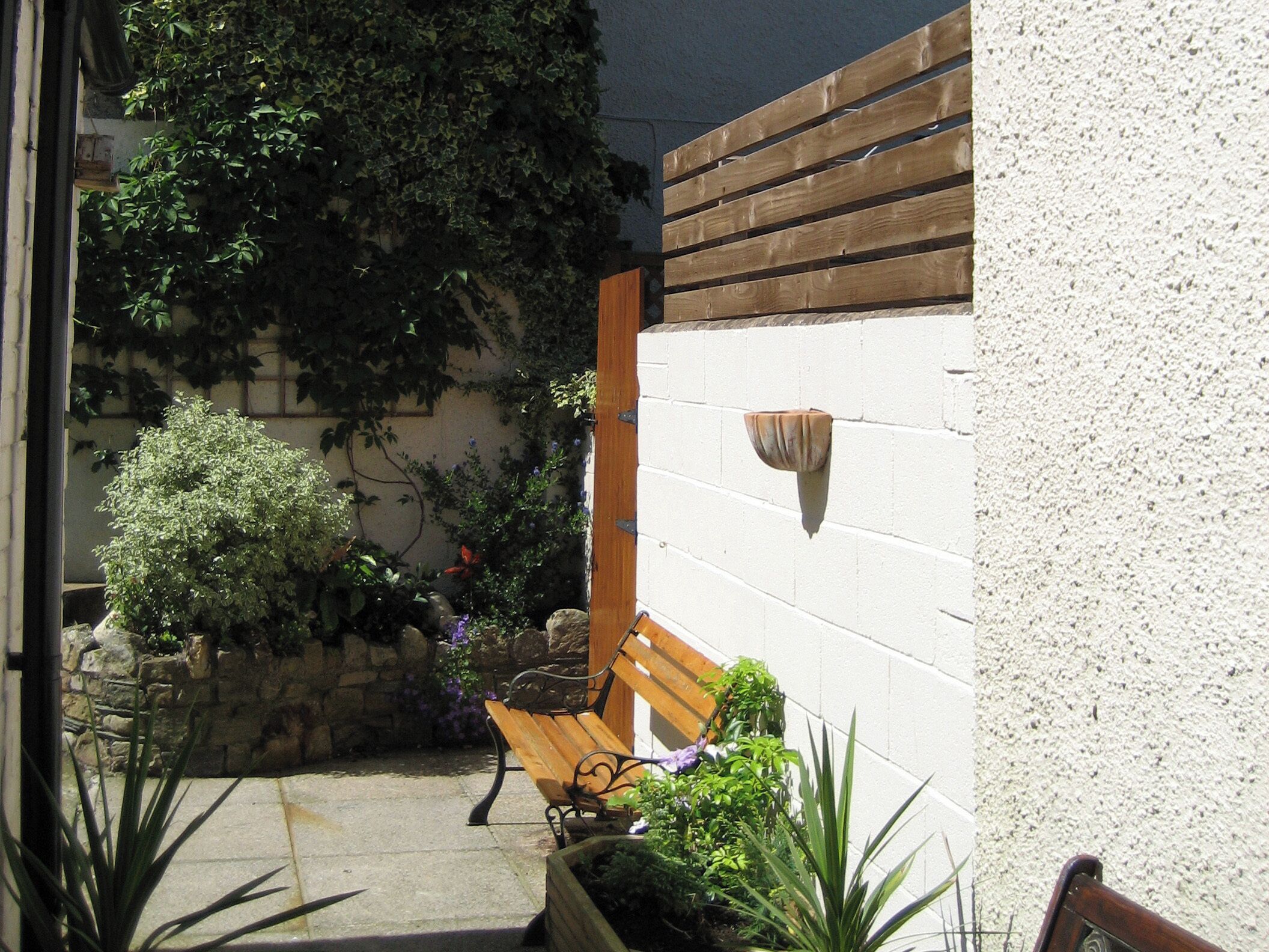 A small outdoor patio area with a wooden bench, potted plants, and a white wall with a wooden slat.
