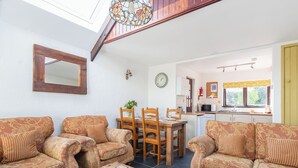 A cozy living room with a wooden coffee table, two patterned sofas, and a dining area with wooden chairs and a table. A kitchen with a window and a yellow curtain is visible in the background. - 2 bedroom accommodation in St Breward, Bodmin Moor (St Breward, Bodmin Moor)