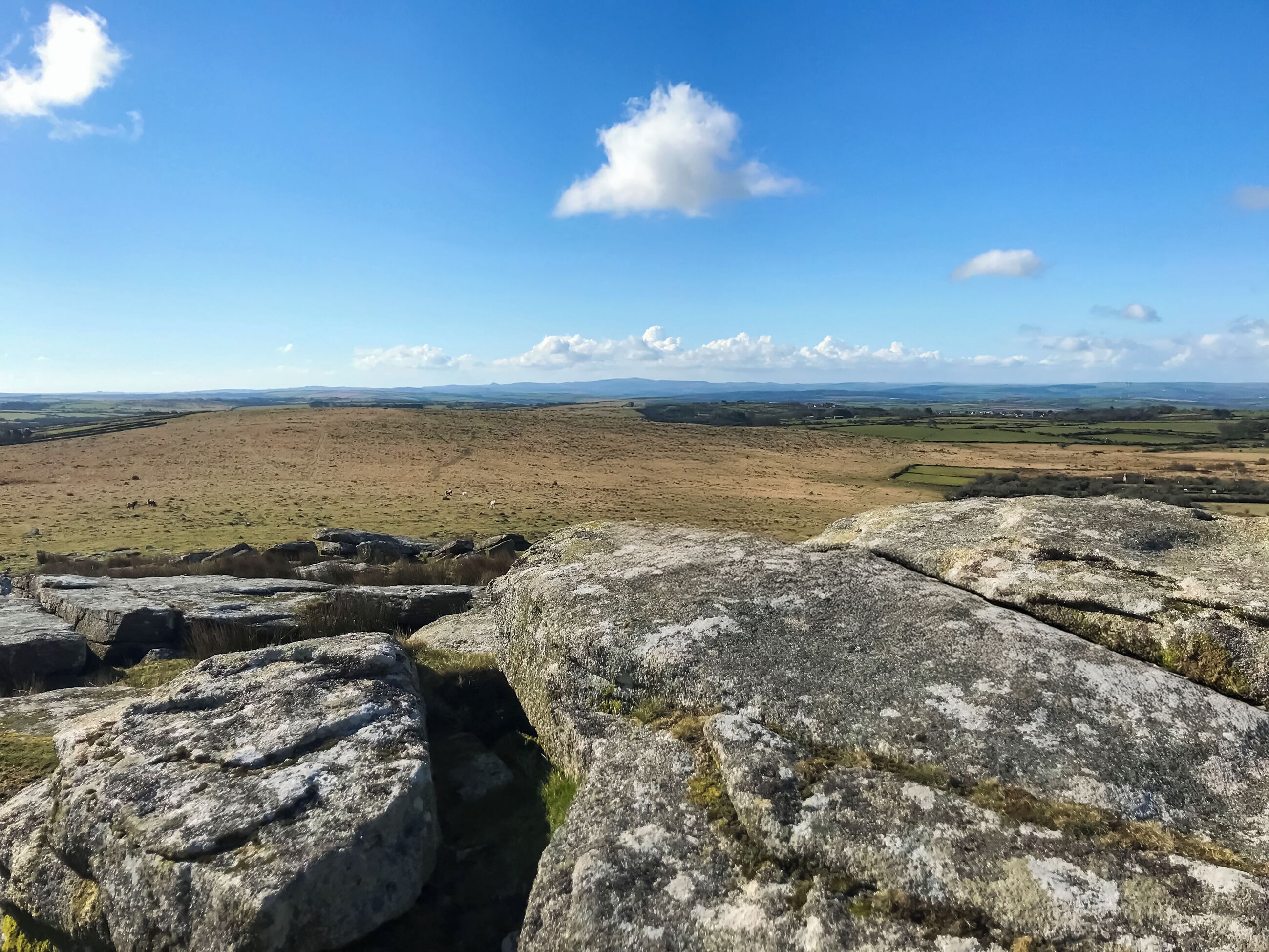A rocky landscape with a distant view of fields and hills under a clear sky.
