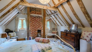 A cozy attic bedroom with exposed wooden beams, a brick fireplace, a bed with floral bedding, and a television on a wooden stand. - 1 bedroom accommodation in Brockdish, near Harleston (Brockdish, near Harleston)