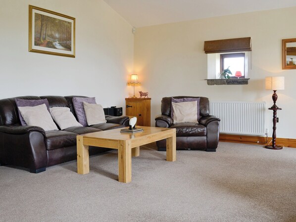 A living room with a dark leather sofa, a wooden coffee table, and a matching armchair. There is a framed painting on the wall, a lamp, and a radiator. - Ground Floor:
Open plan living space. (Silsden near Skipton)