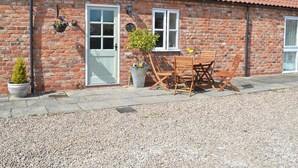 A brick building with a tiled roof, a white door, a window, and a table with chairs outside.