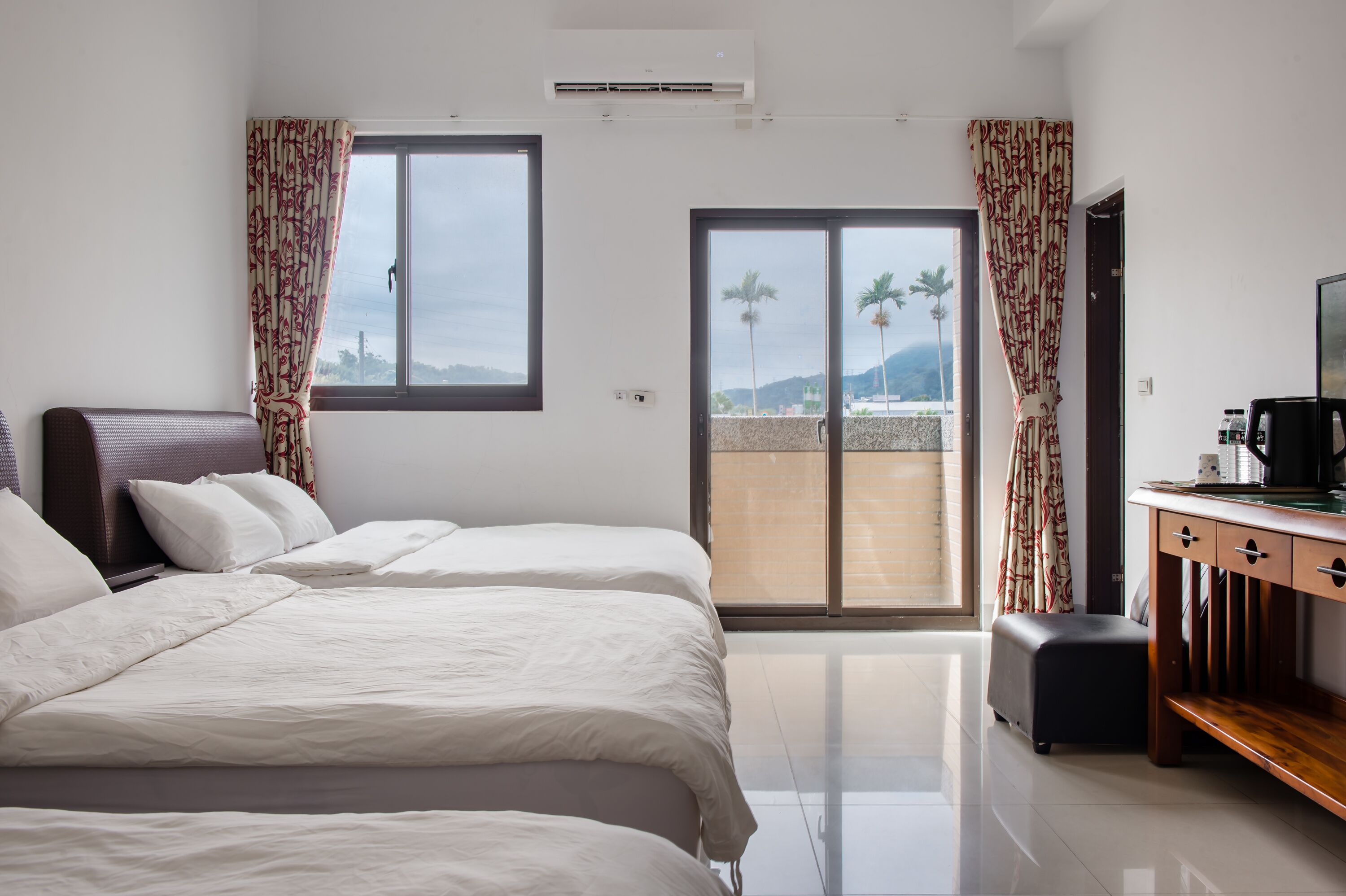 A hotel room with a large bed, a wooden desk, a television, and a sliding glass door leading to a balcony with a view of palm trees and mountains.