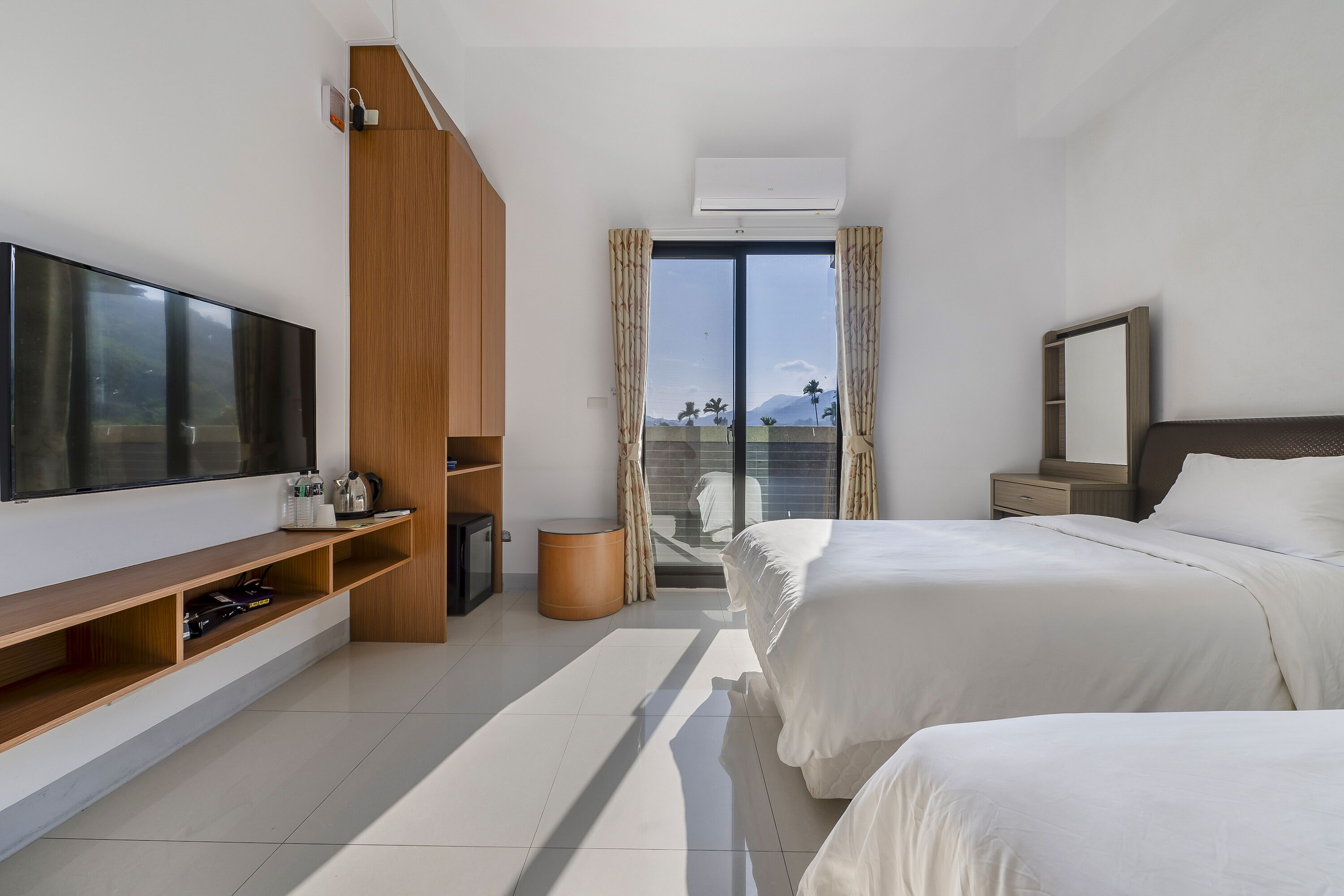 A modern hotel room with a large flat-screen TV, a wooden shelf with amenities, a bed with white linens, a bedside table, and a view of palm trees through a glass door.