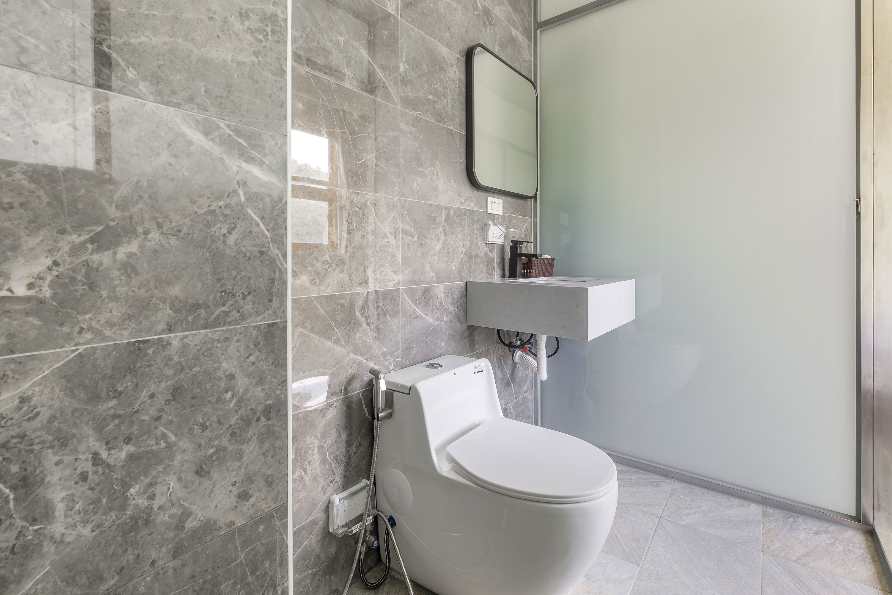 Un baño moderno con inodoro blanco, pared de azulejos de mármol, cabina de ducha de vidrio y lavamanos blanco.