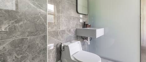 Un baño moderno con inodoro blanco, pared de azulejos de mármol, cabina de ducha de vidrio y lavamanos blanco.