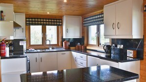 A kitchen with wooden cabinets, a black countertop, and a window with a checkered valance. - 2 bedroom accommodation in Fritton, near Great Yarmouth (Fritton, near Great Yarmouth)