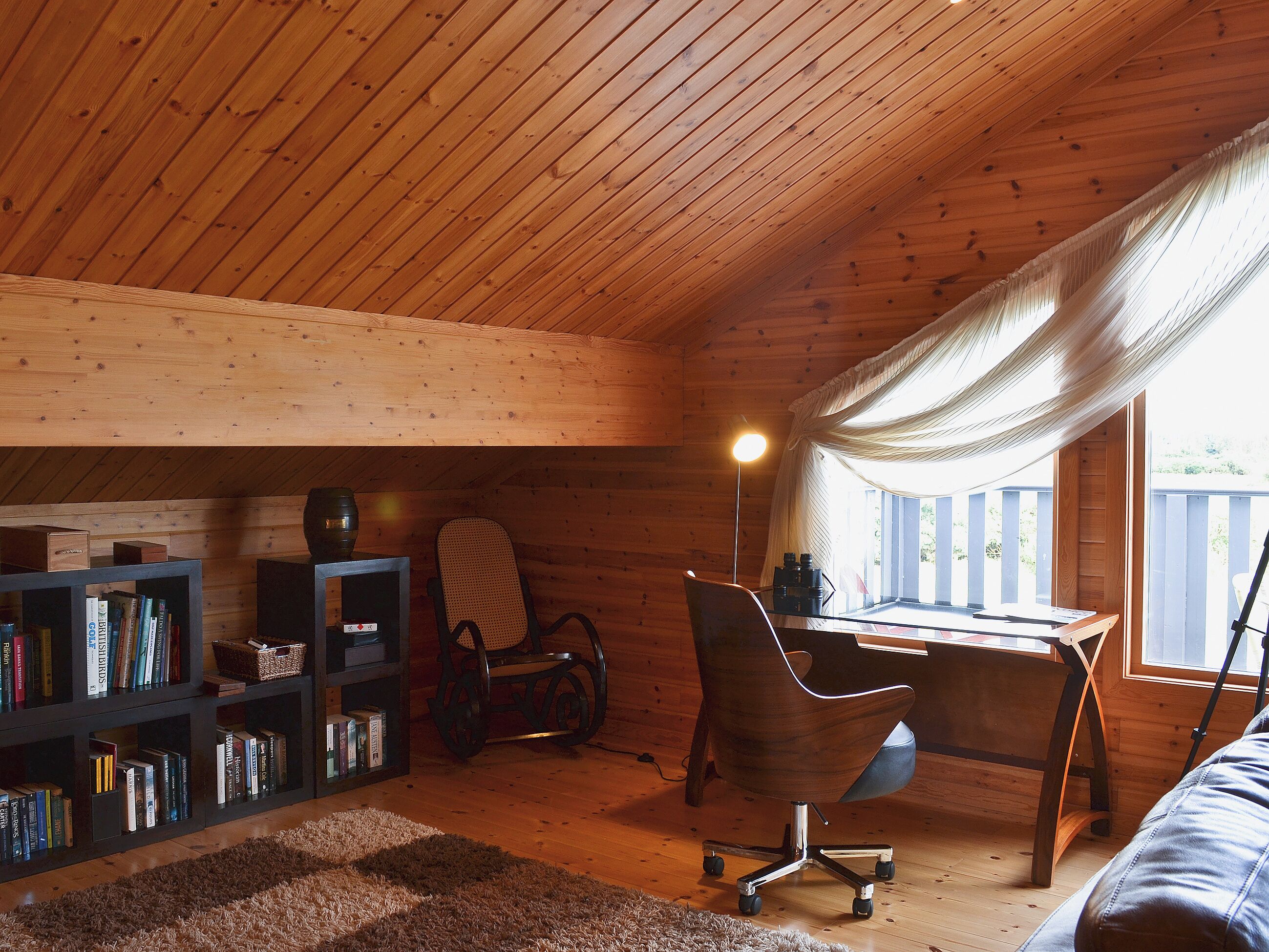 L’intérieur d’un chalet accueillant, avec un plafond en bois, un bureau avec une chaise, une bibliothèque et une fenêtre avec des rideaux.