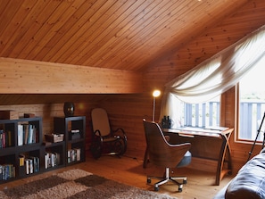 A cozy cabin interior with a wooden ceiling, a desk with a chair, a bookshelf, and a window with curtains.