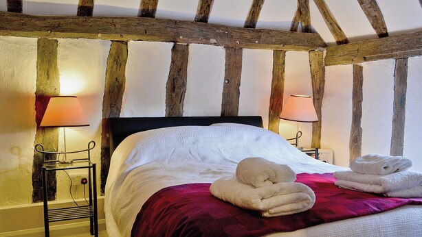 Une chambre avec des poutres apparentes, un lit recouvert de linge de lit blanc, deux lampes de chevet et une serviette pliée posée sur le lit.