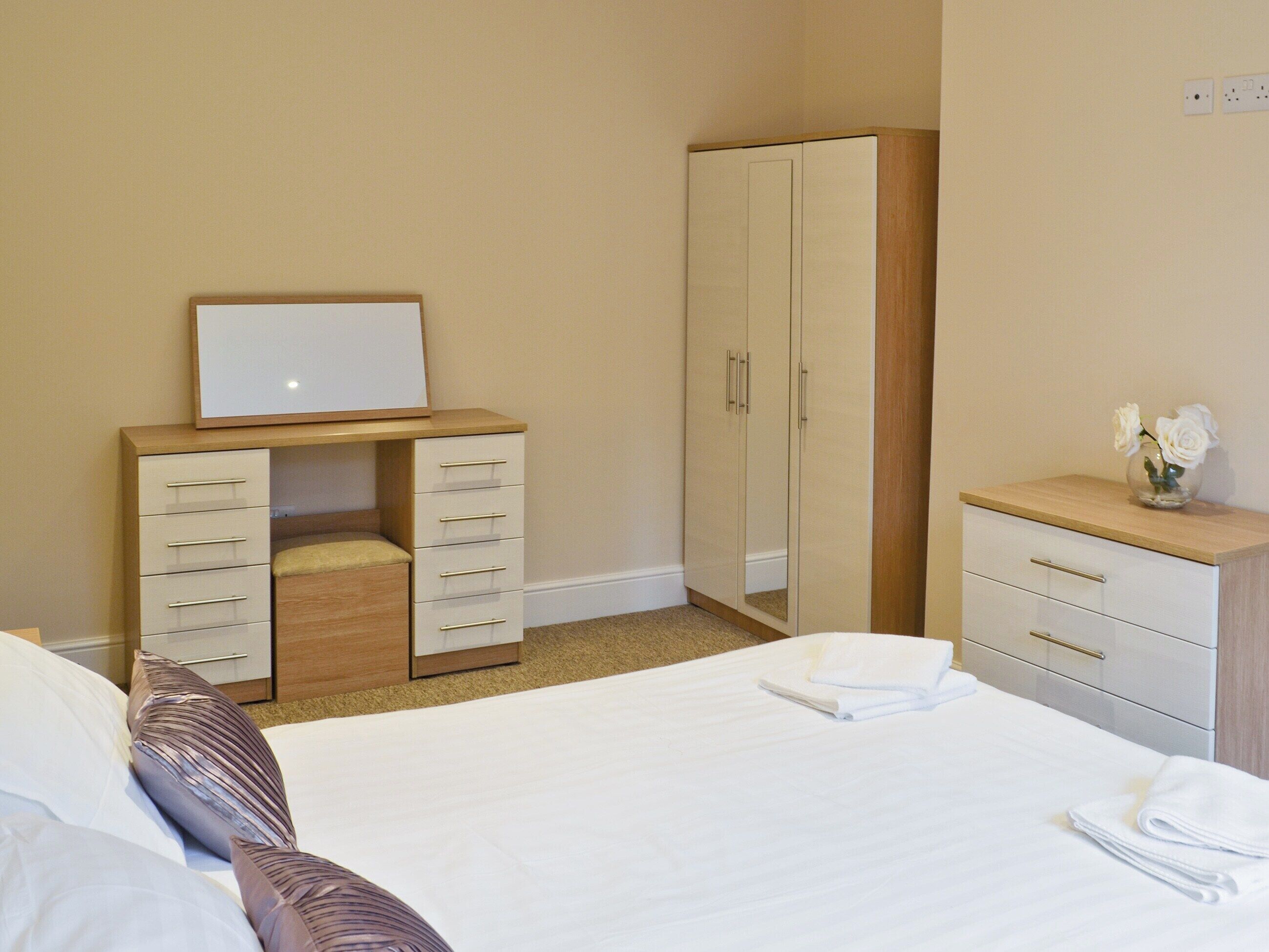A hotel room with a bed, a wardrobe, a desk with a mirror, and a small table with a vase of flowers.