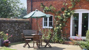 A patio with a wooden bench and table, a green umbrella, and a brick building with a window and door. - 2 bedroom accommodation in Stanhoe, near King&rsquo;s Lynn (Stanhoe, near King&rsquo;s Lynn)