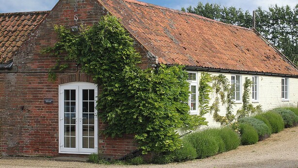 Une petite maison en briques avec une porte et des fenêtres blanches, entourée de verdure et bordée d’un chemin de gravier.