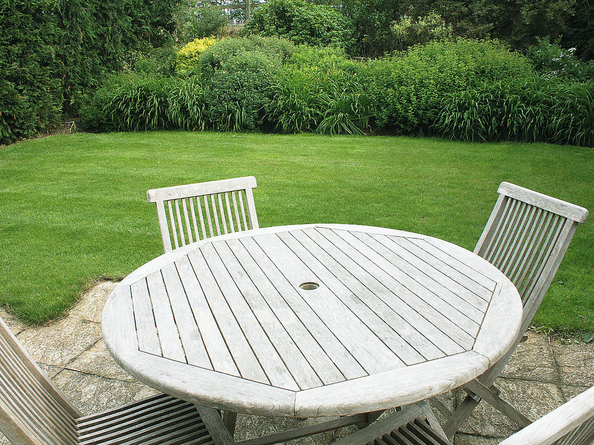 Une table ronde en bois, entourée de quatre chaises, est placée sur une terrasse en pierre. On aperçoit, en arrière-plan, une pelouse bien entretenue et une végétation dense.