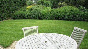 Une table ronde en bois, entourée de quatre chaises, est placée sur une terrasse en pierre. On aperçoit, en arrière-plan, une pelouse bien entretenue et une végétation dense.