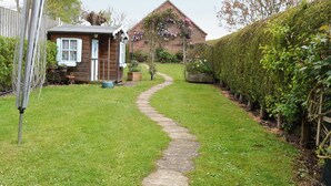 A garden path leading to a wooden cottage with a brick house in the background.