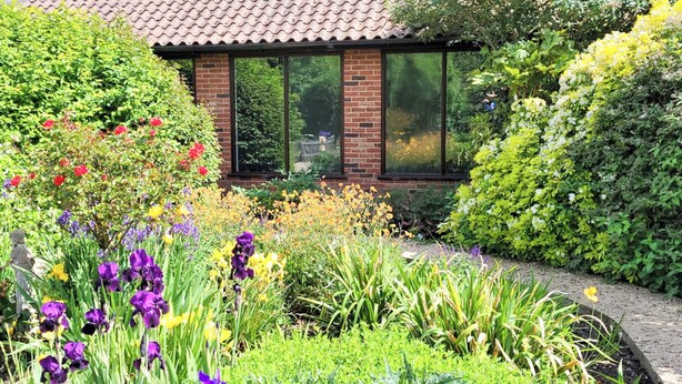 Un jardín con diversas flores y plantas, un edificio de ladrillo con ventanas amplias y un techo de tejas.