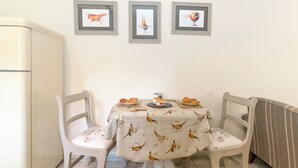 Un comedor con una mesa dispuesta para dos personas, sillas, un refrigerador y tres cuadros en la pared.