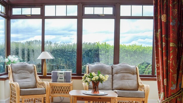 A room with wicker furniture, a table with a vase, and large windows with a view of greenery.