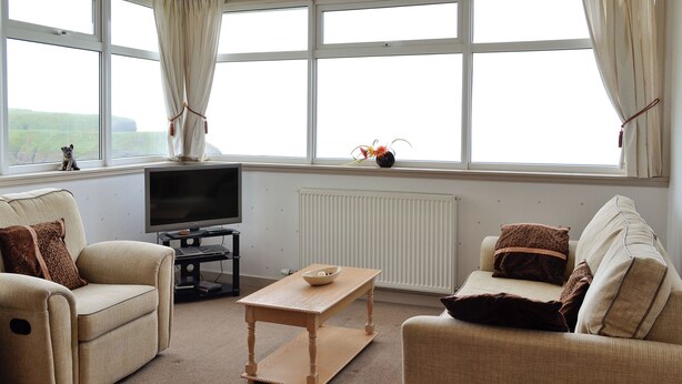 A living room with two sofas, a coffee table, a TV on a stand, and large windows with curtains. - MILL SHORE COTTAGE - 26817 (Pennan, near New Aberdour)