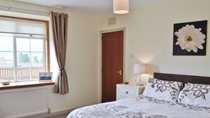 A bedroom with a large bed, a bedside table with a lamp, a window with curtains, and a door. - MILL SHORE COTTAGE - 26817 (Pennan, near New Aberdour)