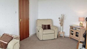 A living room with a beige armchair, a small side table with a lamp, and a wooden cabinet. - MILL SHORE COTTAGE - 26817 (Pennan, near New Aberdour)