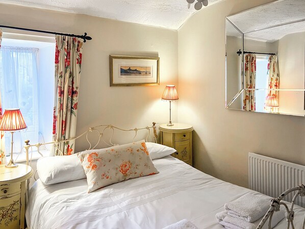 Una cama bien tendida con almohadas estampadas, una lámpara de noche y una mesita de noche. Se ve una ventana con cortinas, un cuadro en la pared y un radiador.