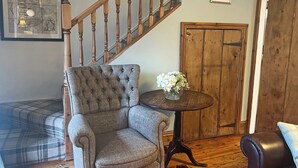 Una sala de estar con un sillón capitoné, una escalera de madera, una puerta de madera y una pequeña mesa con flores.