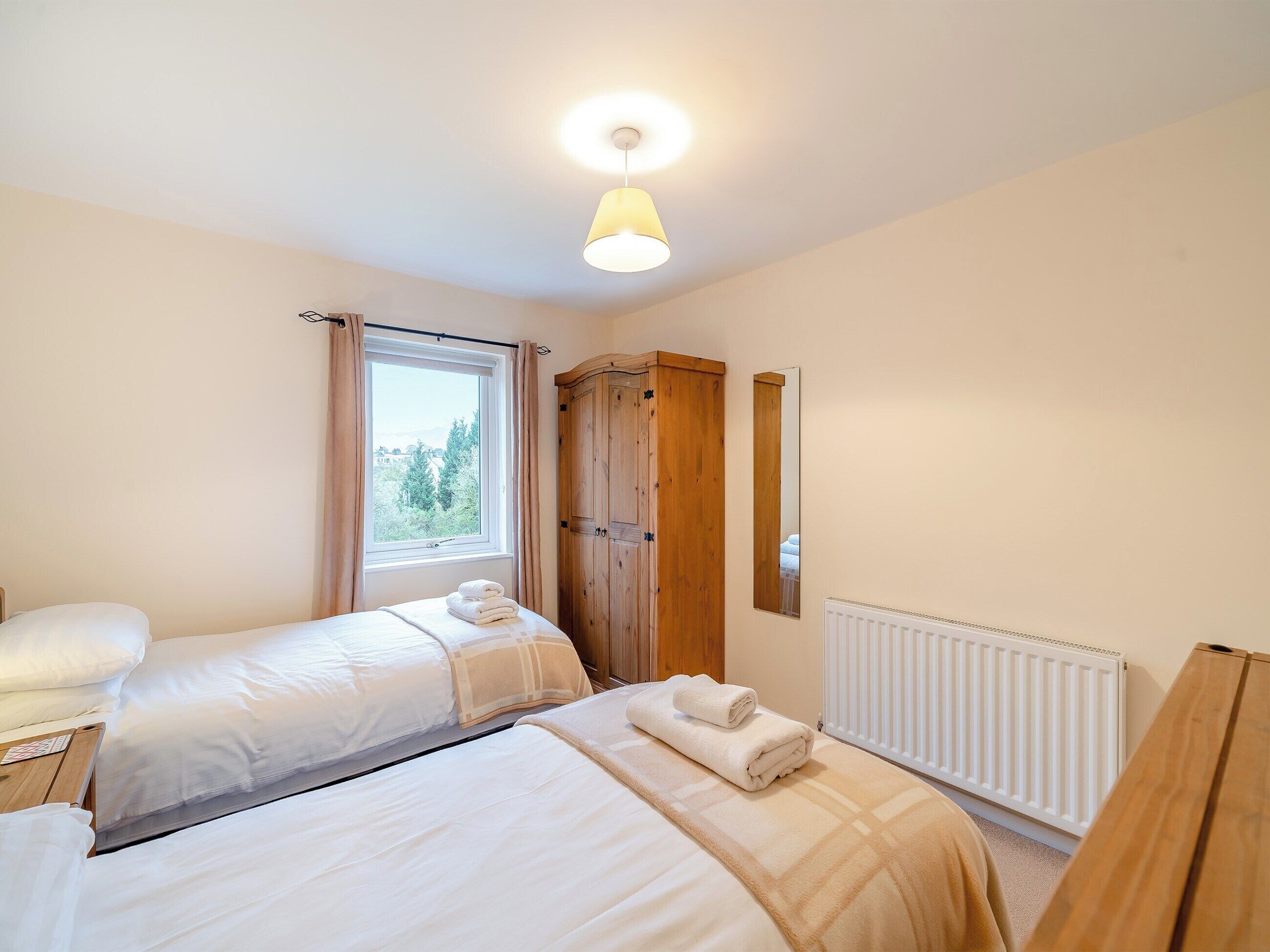 Een slaapkamer met twee bedden, een raam met gordijnen, een houten kledingkast, een radiator en een hanglamp.