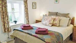 A neatly made bed with a colorful quilt, a bedside table with a lamp, and a window with curtains. - Meadow Cottage-E1793 (Broadwindsor)