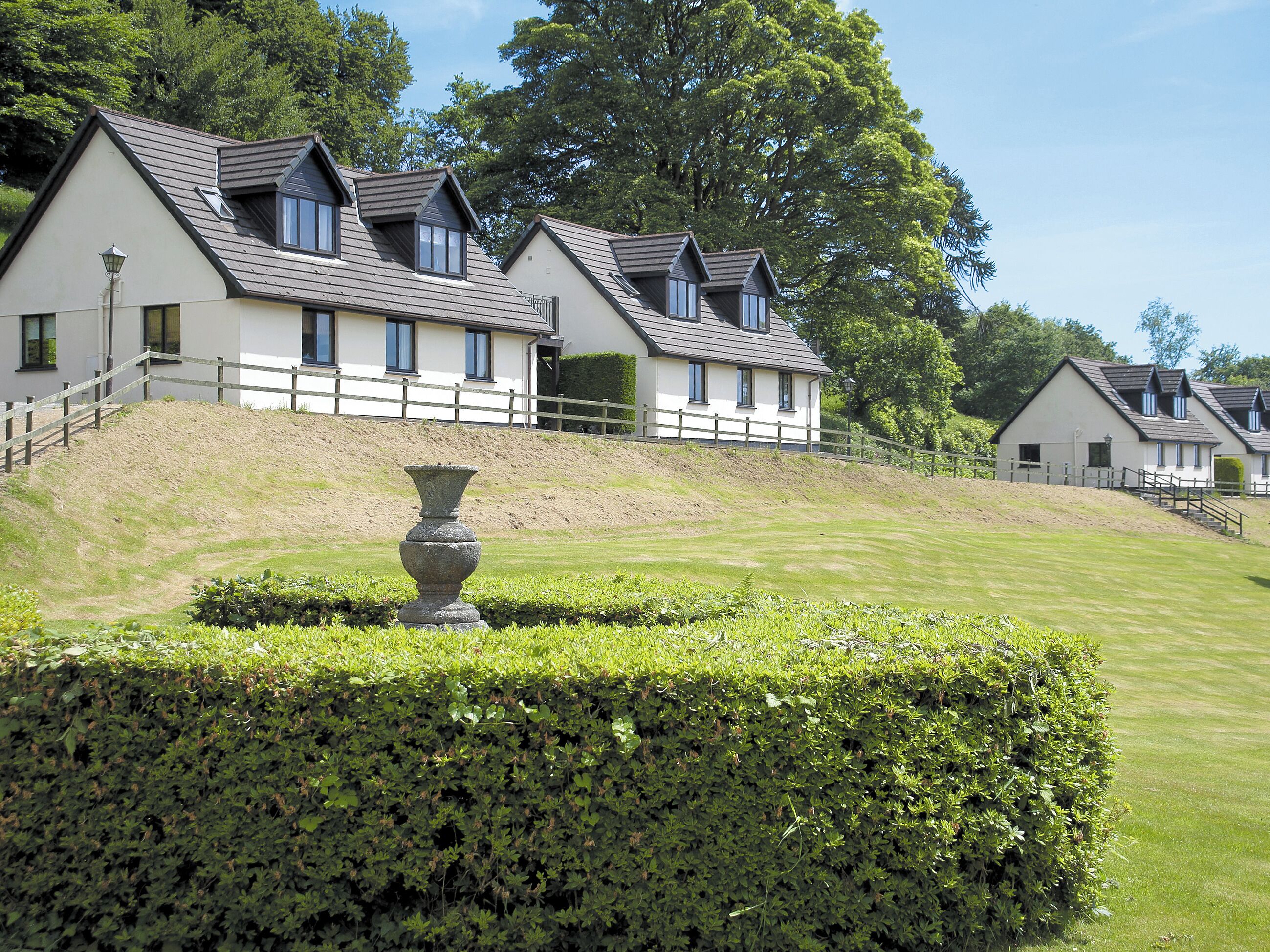 A row of white buildings with grey roofs, a stone urn, and well-maintained hedges.