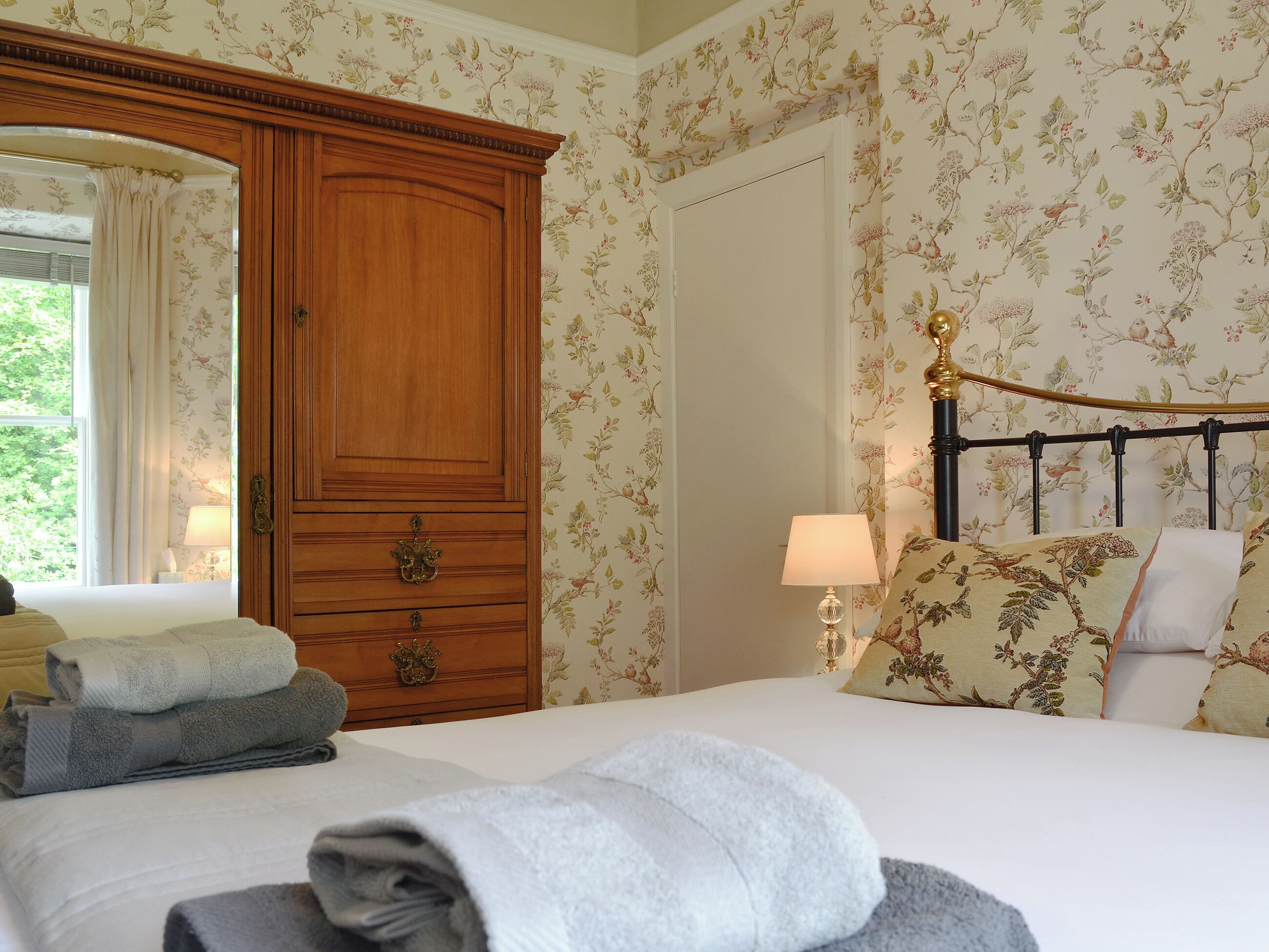 A bedroom with a wooden wardrobe, a bed with floral-patterned bedding, and a window with curtains.