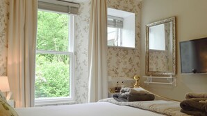 Ein Schlafzimmer mit großem Fenster, einem Bett mit weißer Bettdecke, einem goldfarbenen Spiegel und einem wandmontierten Flachbildfernseher.