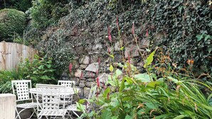 Une terrasse avec des chaises et une table en métal blanc, entourée d’une végétation luxuriante et d’un mur de pierre.