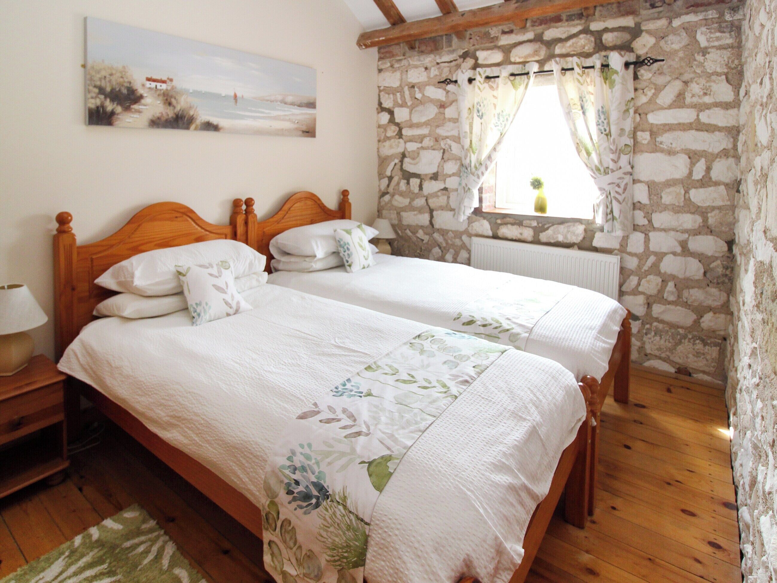A bedroom with two single beds, a wooden headboard, a stone wall, a window with curtains, and a painting on the wall.
